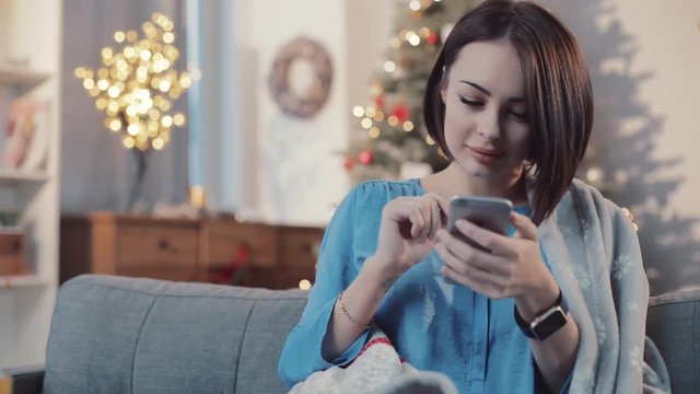 Charming smiling woman drinks tea use pnone sit on the sofa in home background christmas tree apartment brunette girl home mobile internet cell cellphone communication online slow motion