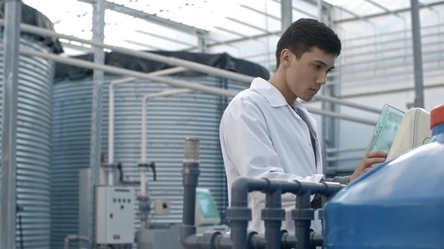 PAN of young male agrochemist in lab coat operating fertigation machine in industrial greenhouse with hydroponic system
