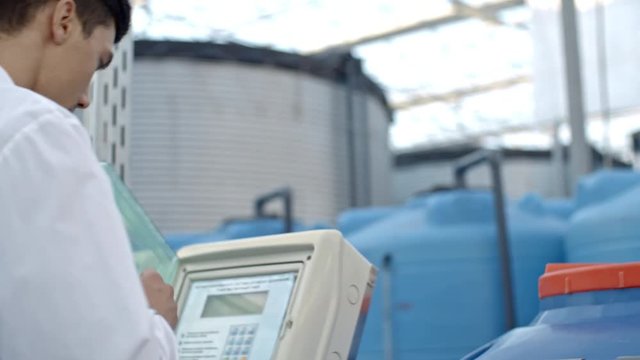 PAN of young male agronomist in lab coat operating fertigation machine in industrial greenhouse with hydroponic system