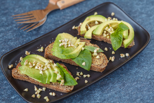 Avocado sandwich with basil and sprouts of green buckwheat on a black straight-headed plate, dark blue background