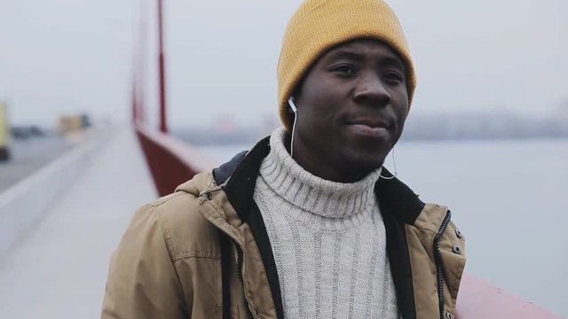Young black smiling man portrait at cold winter standing on a bridge listen earphones