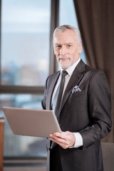 Serious work. Jolly joyful senior man posing on blurryd backgrounds while has tablet and smiling to the camera 