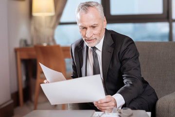 Documents check. Skillful senior appealing businessman working with documents and staring at them while relaxing in the armchair