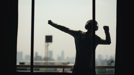 Fototapeta premium Silhouette of young man dancing ad listening music in wireles headphones stand on hotel room balcony