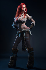 Beautiful red haired woman in stone age clothing with spear posing at camera on dark background