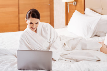 Morning check. Energetic beautiful cheerful woman looking at the screen while relaxing on the bed and wearing bathrobe 