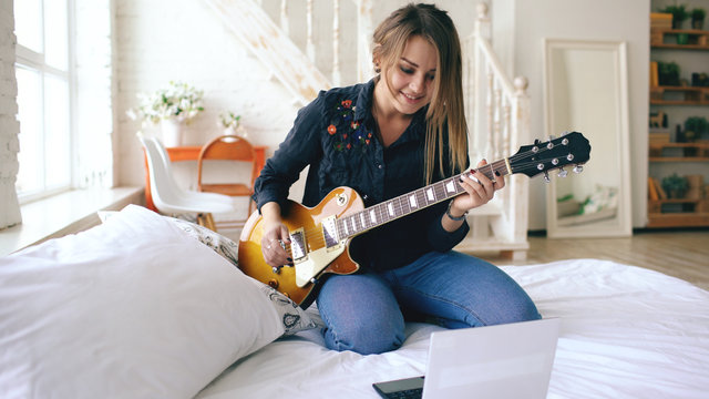 Attractive young girl learning to play electric guitar with notebook sit on bed in bedroom at home