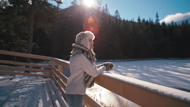 Slender Young Lady is Admiring the Mountain Lake under snow in Winter with Amazing Sun Lense Flare