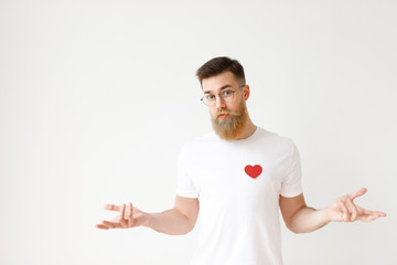 Uncertain hesitant bearded male in casual t shirt shruggs shoulders in bewilderment, tries to make decision about future life, isolated over white studio background. Doubtful man poses indoor