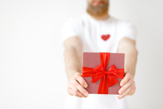 Cropped image of unrecognizable bearded male holds wrapped present box with red ribbon, isolated over white studio background, going to present it to girlfriend. Gift for you. Holiday concept
