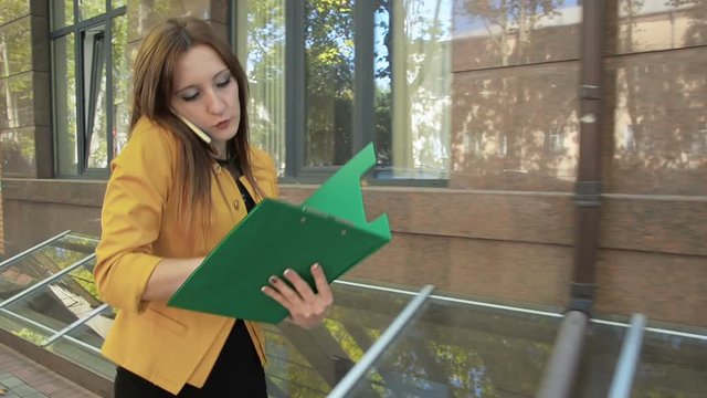 Woman walks down street and talks on phone and looks through important financial documents. Business woman with green folder goes to business district