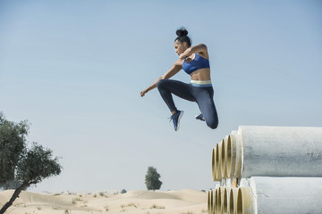 Black African Amercian athletic woman jumps over and leaps from construction pipes wearing sports outfit in a parkour or extreme fitness competition wearing a sports outfit. 