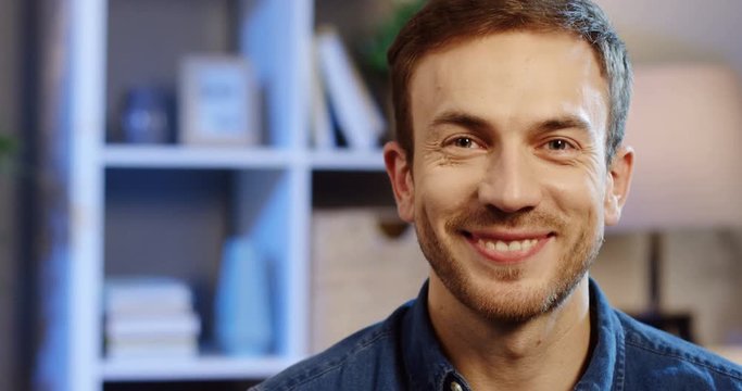 Portrait of the handsome caucasian man smoling joyfully in front of the camera in the living room at night. Close up. Inside