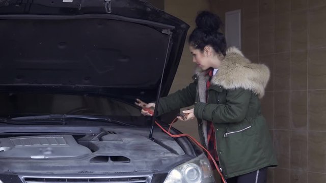 Beautiful woman with jumper cables near car in garage