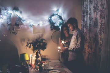 happy new year and merry christmas concept. happy stylish hipster couple holding burning sparklers fireworks and embracing in room at hoome in evening. happy holidays. toned