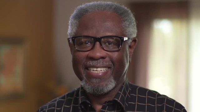Elderly African American male with glasses smiles in a close up authentic portrait, cheerful and happy, looking at camera. Prores file.