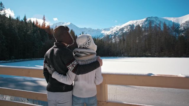 Young Happy Couple is Standing near Mountain Lake in Sunny and Snowy Winter Looking at Amazing Landskape