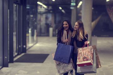 Two cute young adolescent women, heavy loaded with christmas gifts,  shopping, while walking along store windows and choosing and admiring the items. 