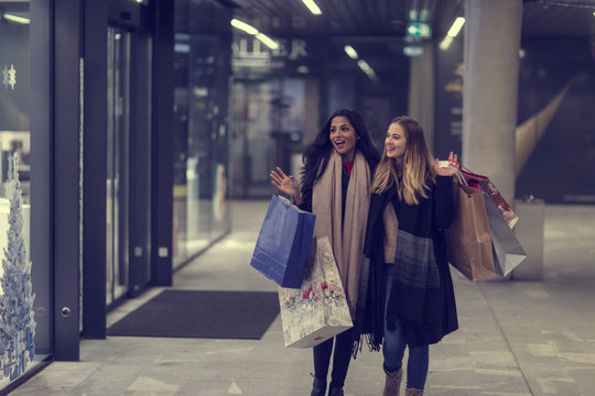 Two cute young adolescent women, heavy loaded with christmas gifts,  shopping, while walking along store windows and choosing and admiring the items. 