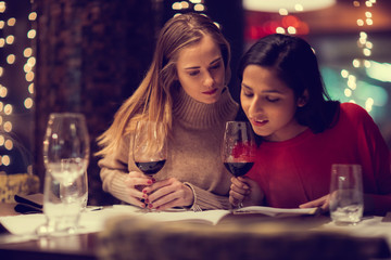 Two adolescent women going through the menue  in a fancy restaurant, while drinking a glass of red wine and laughing and talking, maybe gossiping