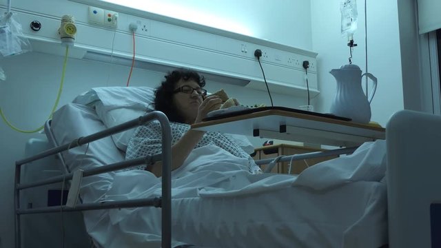 Middle aged woman with glasses, patient in hospital gown is sitting in profiling bed and having a sandwich 