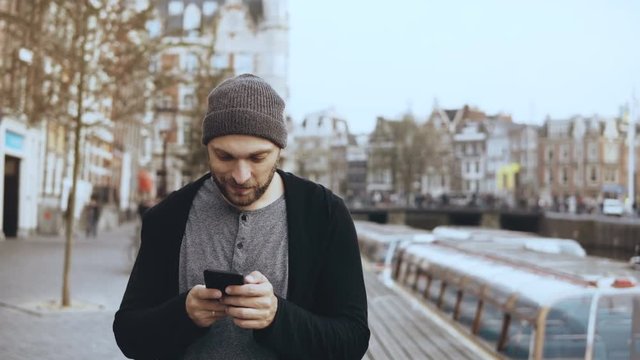 4K Caucasian tourist man walking with smartphone. Casual handsome bearded smiling adult male texting in the street.