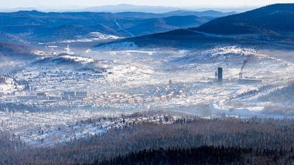 View of a small village Sheregesh, high mountains and coniferous forest from a high point in winter on a clear day with blue sky and fog