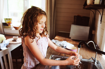 child girl helps mother at home and wash dishes in kitchen. Casual lifestyle in real interior. Teaching kids to do house work.