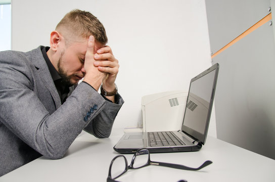 a tired man with his hands on the his face at the table with a laptop in the office