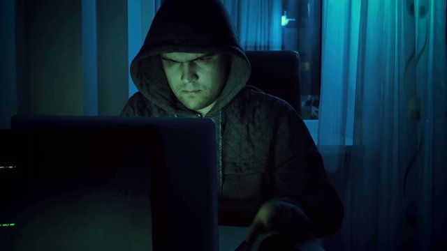 4k footage of young male hacker in hoodie working on laptop at home in night. Concept of cyber crime