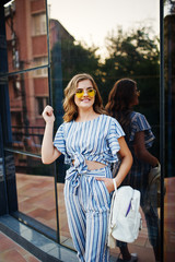 Portrait of a gorgeous young woman in striped overall and yellow sunglasses posing with white leather backpack on a balcony of a modern glass building in town.