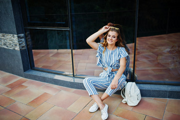 Portrait of a fabulous young woman in striped overall and sunglasses sitting on the ground and posing with a backpack against glass wall.