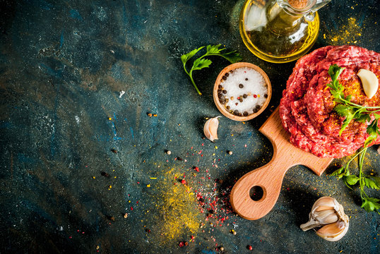 Raw burger cutlets with salt, pepper, oil, herbs and spice, on dark table, copy space top view