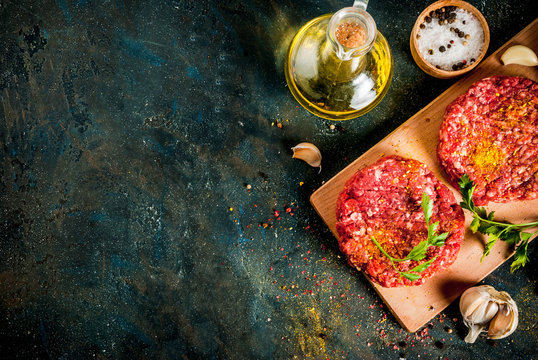 Raw burger cutlets with salt, pepper, oil, herbs and spice, on dark table, copy space top view