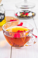 A cup of tea, in the background of a bank of honey and a jar with a black herbal floral tea on a white wooden table.