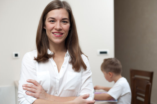 Concept Of Happy Mother. Young Positive Woman In White Shirt Smiling And Looking At Camera On Background Of Drawing Child