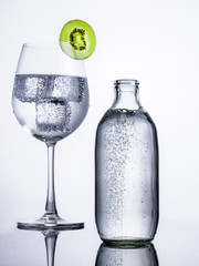 A glass of soda water with ice and a sliced kiwi fruit and a bottle of soda water on a reflected floor and bright background