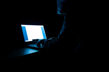 Credit card thief wearing hood, sitting in the front of laptop, credit cards, in a dark room,...