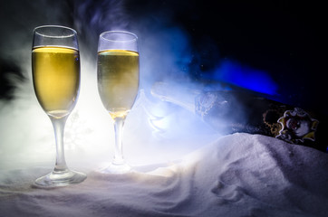 New Year Eve celebration background with pair of flutes and bottle of champagne with Christmas attributes (or elements) on snowy dark toned foggy background. Selective focus.