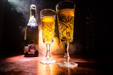 New Year Eve celebration background with pair of flutes and bottle of champagne with Christmas attributes (or elements) on snowy dark toned foggy background. Selective focus.