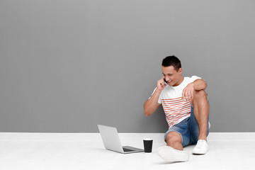 Young man with laptop and mobile phone sitting on floor near grey wall
