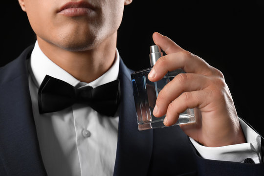 Handsome young man in suit using perfume on black background
