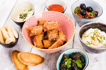 Assortment of appetizers on a white wooden background. Spring rolls with sauce, olives, toast, bread, dip, cheese, olives and sun dried tomatoes in colorful bowls. Party food or finger food.