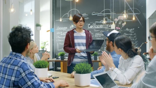 Charismatic Asian Team Leader Shows Laptop to a Diverse Group of Talented Young Developers, They Start Discussion at the Meeting Table. Creative People in Stylish Office Environment. 