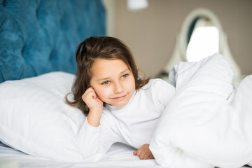 Close-up portrait of beautiful little kid lying on bed with hand under pillow, looking at camera