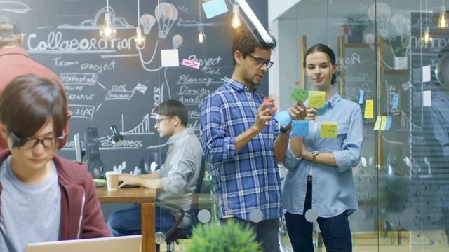 Long Shot of The Busy Office Where Young Creative People Work on Laptops, Personal Computers and Outline Project Plan with Sticky Papers on a Glass Wall. Shot on RED EPIC-W 8K Helium Cinema Camera.