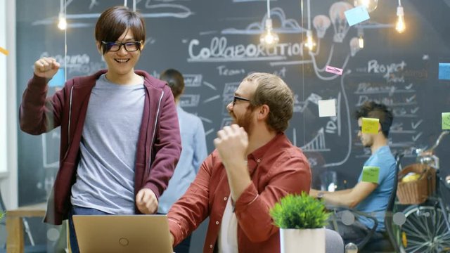 Two Creative Designers Look at the Laptop and Have Great Success, They do High Five and Laugh. In the Background Stylish Office with Talented People Working. 