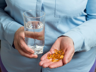 Yellow medication capsules of omega 3, fish oil, healthy supplement pills in the woman palm hand and glass with water in the other hand