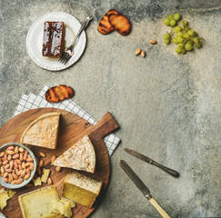 Flat-lay of cheese platter with cheese assortment, green grapes, bread, honey and nuts over grey concrete background, top view, copy space. Party or gathering eating concept
