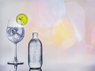 A glass of soda water with ice and a sliced lemon and a bottle of soda water on a reflected floor and multi color bright background